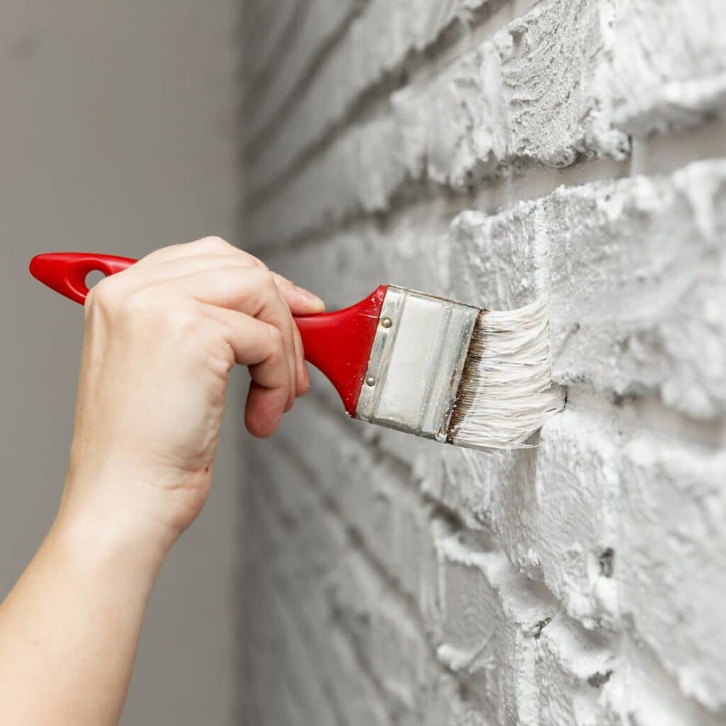 person using a brush to apply white paint to a brick wall