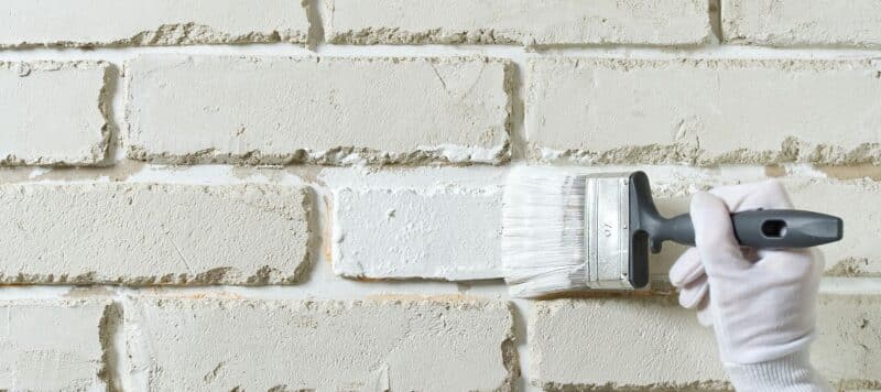 person using a brush to paint a brick wall of a home white