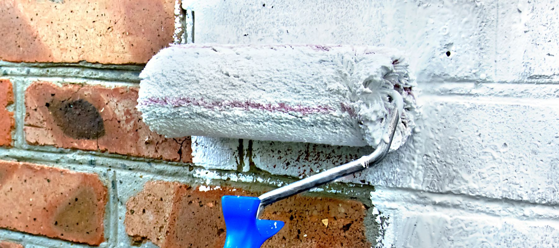 blue paint roller brush spreading white paint on red brick