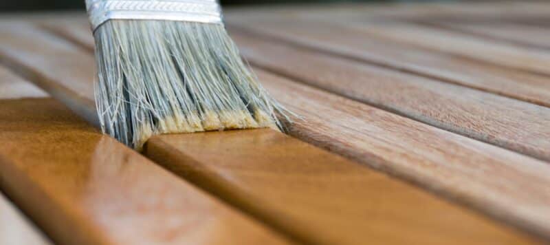 paint brush applying light brown staining liquid to wooden slats on a deck floor