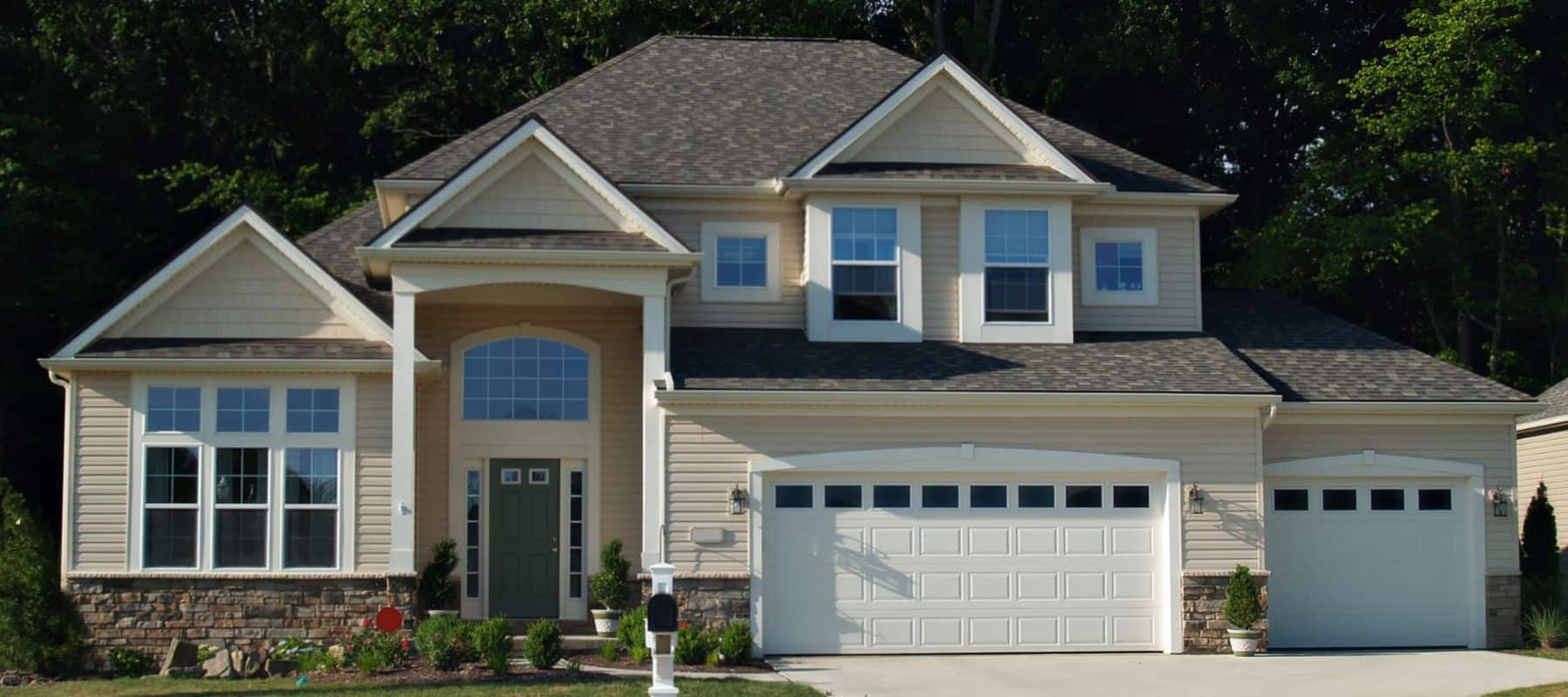Large beige two-story house with multiple garages and manicured landscaping in a residential neighborhood