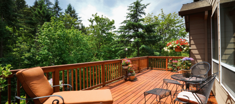 Wooden backyard deck with railing overlooking lush green trees, featuring patio furniture including a cushioned lounge chair, metal table and chairs, and colorful potted flowers in hanging and floor planters.